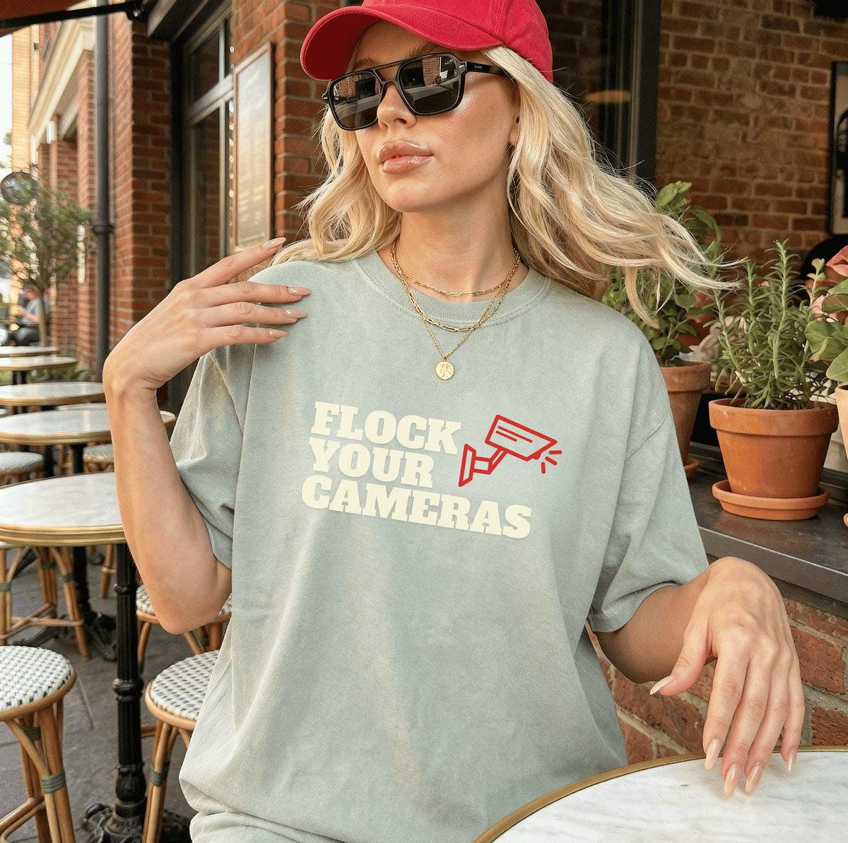 A woman wearing an oversized light sage green graphic t-shirt with the text 'FLOCK YOUR CAMERAS' in cream and red, paired with a red baseball cap and sunglasses, seated at an outdoor cafe table.