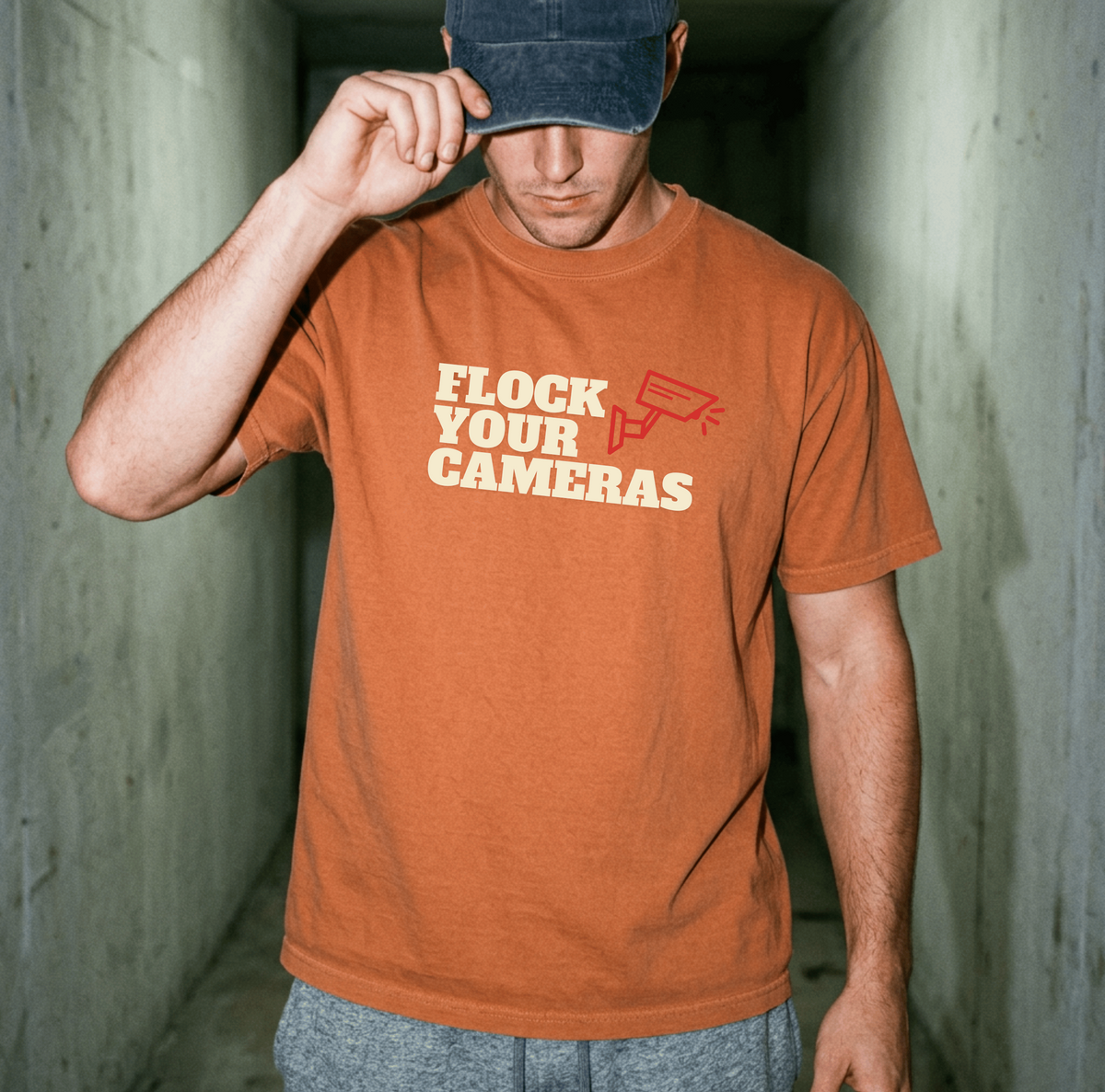 A woman stands on a dock by the water wearing a light blue graphic T-shirt that reads 'FLOCK YOUR CAMERAS' in white and red, with an American flag draped over her shoulders.