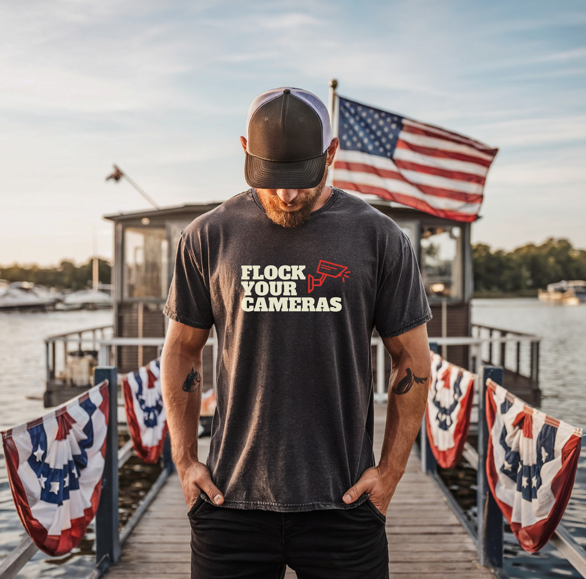 A man wearing a dark charcoal gray t-shirt with a graphic that reads 'FLOCK YOUR CAMERAS' in white and red, standing on a wooden dock with an American flag in the background.